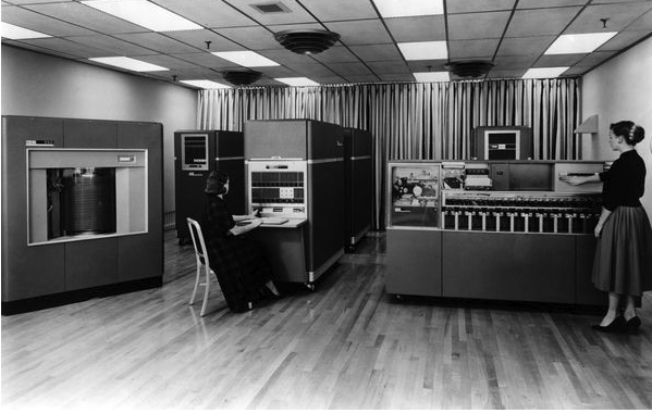Vintage computing room with two people operating mainframe computers from the mid-20th century, showcasing early data processing equipment.
