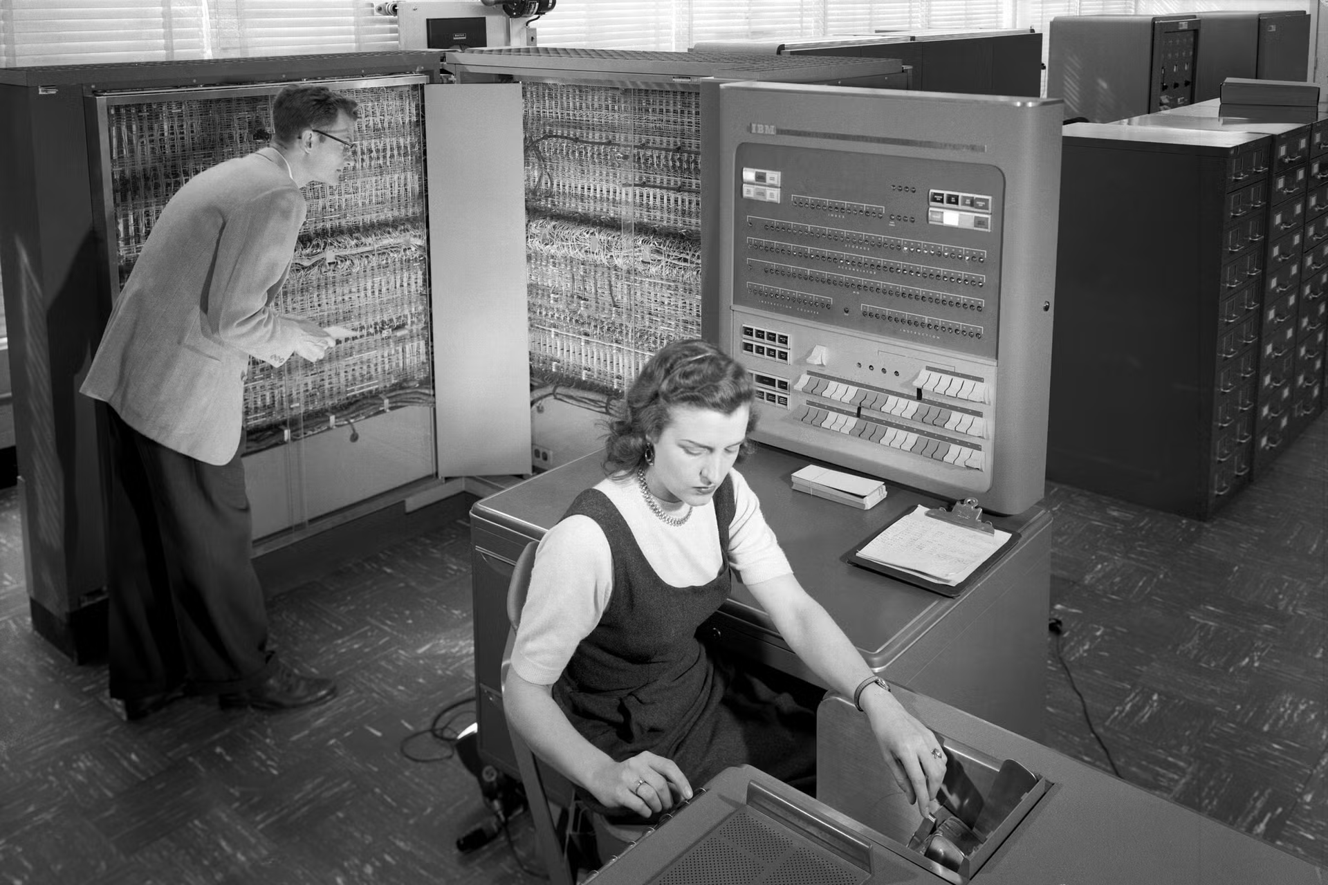 Two professionals operating an early IBM computer system, one working on the database hardware backend while the other inputs data.